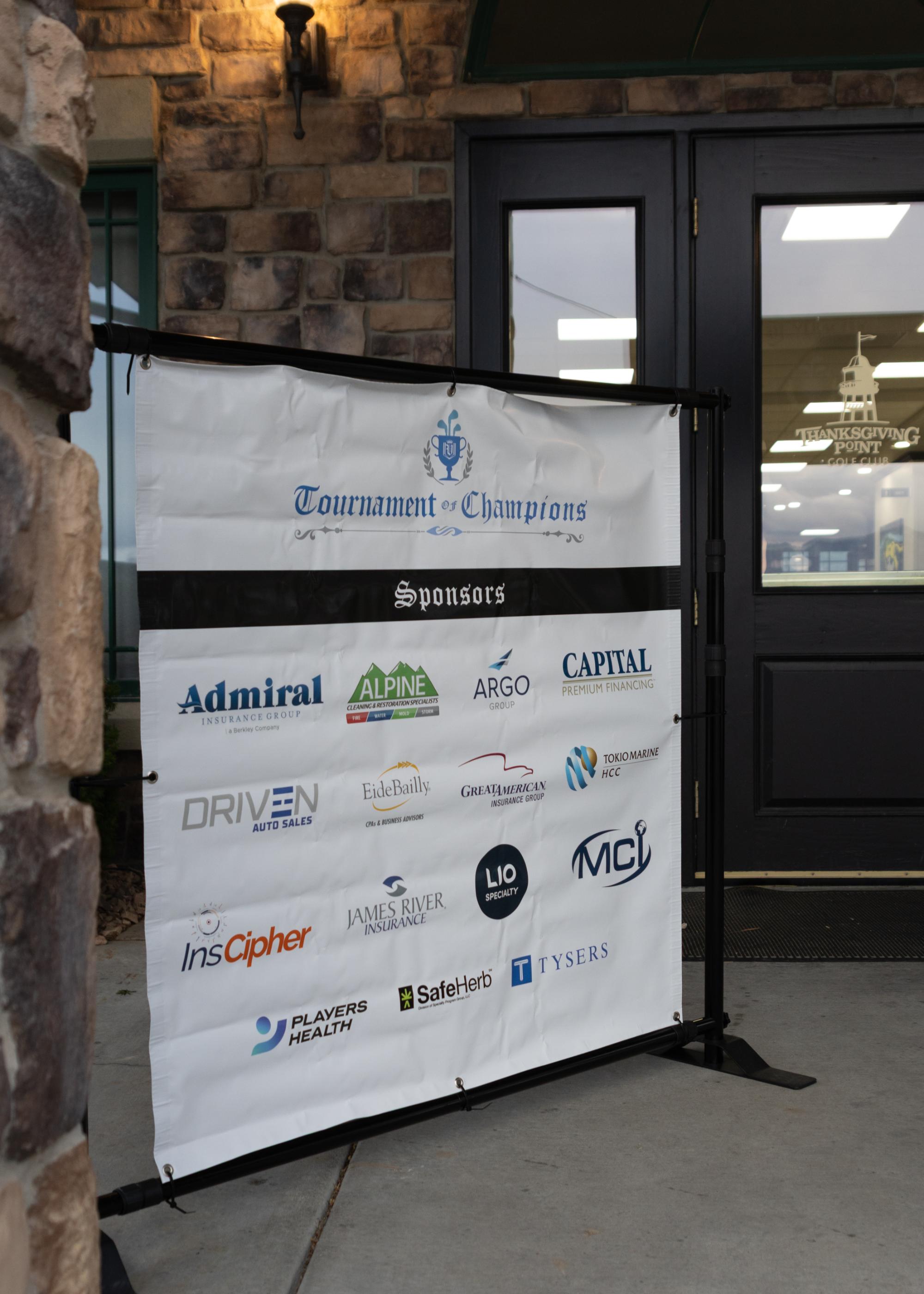 Vinyl banner reading ‘Tournament of Champions’ with dozens of sponsor logos displayed outside clubhouse entrance